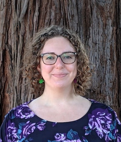 Department photo of Maya, a white woman with curly hair and glasses, with redwood bark in background